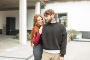 Happy young couple in fashionable urban clothes with hoodie in the city. Cool handsome hipster bearded man with a tattoo and a beautiful girl with red hair with a smile