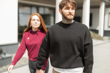 Beautiful young fashionable couple in fashion clothes walks on a sunny day in the city. Handsome hipster man and beautiful red-haired woman in pullover