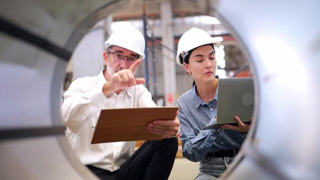 Senior engineer holding a worksheet wearing safety helmet training and explaining to a beautiful female engineer wearing a hard hat, holding a laptop in the factory Industrial steel cutting machine. 