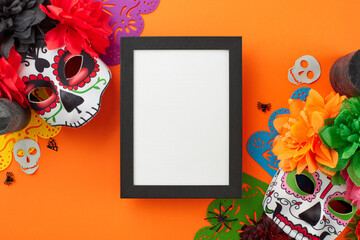 A joyful and enchanting Day of the dead. Top view flat lay of traditional masks with flowers, colorful garland, candles, spooky accents on orange background with empty frame for ad or text