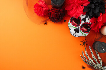 Explore the beauty of elaborate carnival masks and the Day of the Dead spirit. Top view of traditional mask, flowers, candles, skeleton hand, creepy decor on orange background with advert area