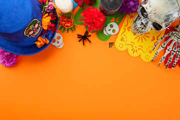 Tradition and celebration of Dia de los Muertos. Top view shot of traditional hat, skull, elaborate flowers, colorful garland, candle, skeleton arm, scary decor on orange background with promo area