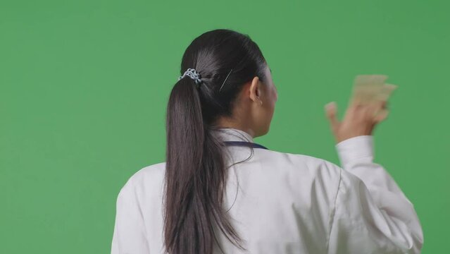 Close Up Back View Of Asian Female Doctor With Stethoscope Waving Hand And Smiling While Walking On Green Screen Background In The Hospital
