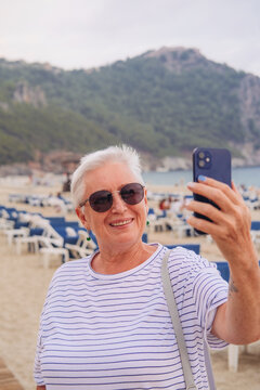 A senior woman with short grey hair, sun glasses, taking a selfie picture on a beach 