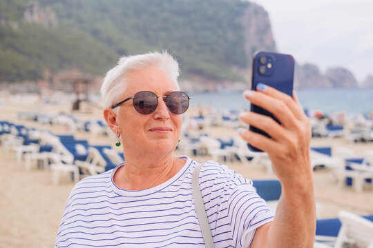 A senior woman with short grey hair, sun glasses, taking a selfie picture on a beach 