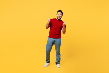 Fototapeta na wymiar Full body young overjoyed happy Indian man wears red t-shirt casual clothes doing winner gesture celebrate clenching fists say yes isolated on plain yellow orange background studio. Lifestyle concept.