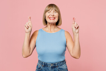 Fototapeta na wymiar Elderly blonde woman 50s years old she wear blue undershirt casual clothes waiting for special moment, keeping fingers crossed, making wish, eyes closed isolated on plain pastel light pink background