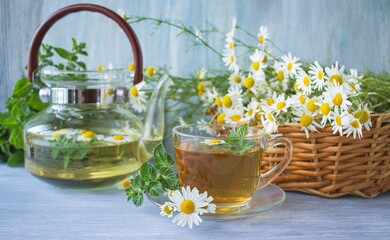 Tea from fresh chamomile flowers, mint, in a cup, on a wooden table, against the background of bouquets of fresh chamomile flowers