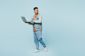 Full body side view young IT man of African American ethnicity wear denim jacket orange t-shirt work hold use laptop pc computer isolated on plain pastel light blue cyan background. Lifestyle concept.