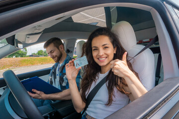 Happy lady holding license and showing thumbs up while male instructor writing on clipboard in car. Driving test, driver courses, exam concept - obrazy, fototapety, plakaty