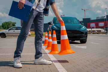 Male driving instructor standing with orange traffic cones and clipboard in his hands during exam outdoors. Driving test, driver courses, exam concept - obrazy, fototapety, plakaty
