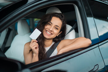 Cheerful young lady sitting in her car and showing her new driving license. Driving test, driver courses, exam concept
