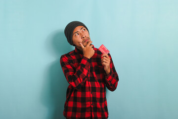 Pensive young Asian man is holding credit cards looking at copy space, isolated on blue background