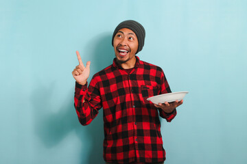 Excited Young Asian man is pointing at copyspace while holding white plate isolated on blue backdrop