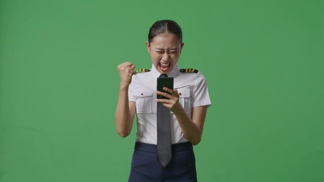 Asian Woman Pilot Looking At Smartphone Then Screaming Goal Celebrating While Standing In The Green Screen Background Studio
