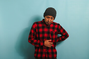 Unhappy young Asian man is suffering from a stomach ache, bending over, isolated on blue background