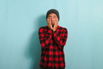 Sleepy bored young Asian man yawns, keeping his hand on his cheeks, isolated on a blue background