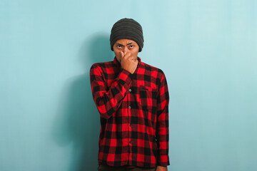 Annoyed young Asian man holds his breath smells something stinky, isolated on blue backdrop - obrazy, fototapety, plakaty
