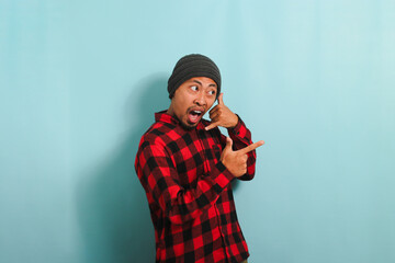 Excited young Asian man makes call me gesture, pointing to the right, isolated on a blue background