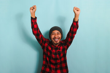Excited young Asian man rejoicing, saying YES with fist pump gesture, isolated on blue background