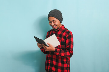 A smiling young Asian man is holding a book while standing against a blue background