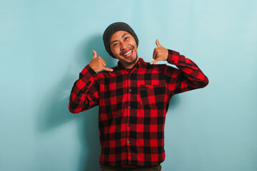 An excited young Asian man makes a CALL ME gesture with his hand, isolated on a blue background