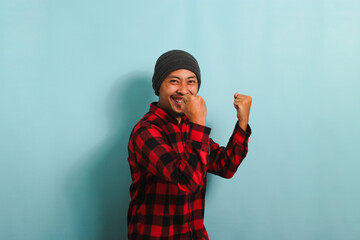 Excited young Asian man rejoicing, saying YES with fist pump gesture, isolated on blue background