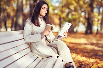 Young  beauty woman sitting with a cup of coffee on a bench in the fall park. A girl reads an e-book in the autumn forest