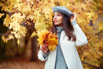 Naklejka na ściany i meble Autumn portrait of a beautiful happy smiling woman with yellow maple leaves in a gray beret in the park