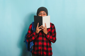 A young Asian man is covering his face with a book while standing against a blue background