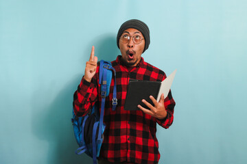 Excited Young Asian student is holding books and pointing while standing against a blue background