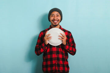 Happy Young Asian man is smiling while holding an empty white plate, isolated on a blue background - obrazy, fototapety, plakaty