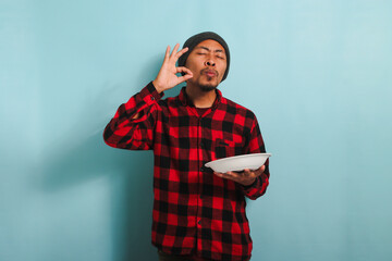 Young Asian man is making delicious hand gesture while holding plate, isolated on blue background