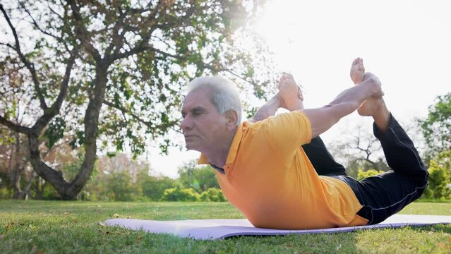 Indian Asian senior citizen man or Fit old male performing a yoga asana called Dhanurasana (bow pose) on a mat in garden against morning sunlight. Healthy lifestyle, health-conscious, exercise concept