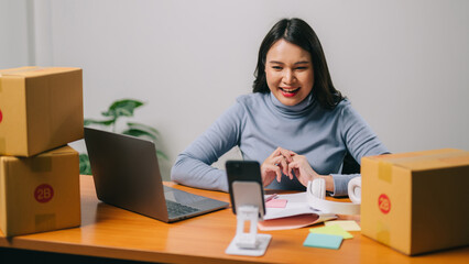 Happy asian woman using mobile phone to live broadcast on social media with parcel box and computer laptop and sitting on chair, Online marketing packing box delivery concept. Online shopping.