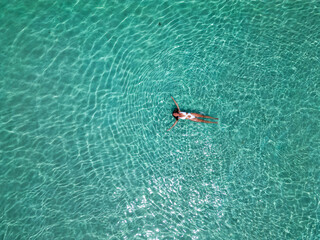 Top view of a young woman in the transparent, blue ocean. Aerial view of a swimming woman lying and floating on the water in the Maldives