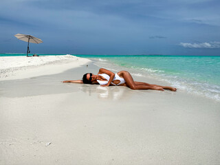 A fit sexy tan brunette girl in a white swimsuit and sunglasses laying on the beach by the Indian Ocean. The shores of the ocean gently lap at the sand, creating a soothing backdrop to her relaxation