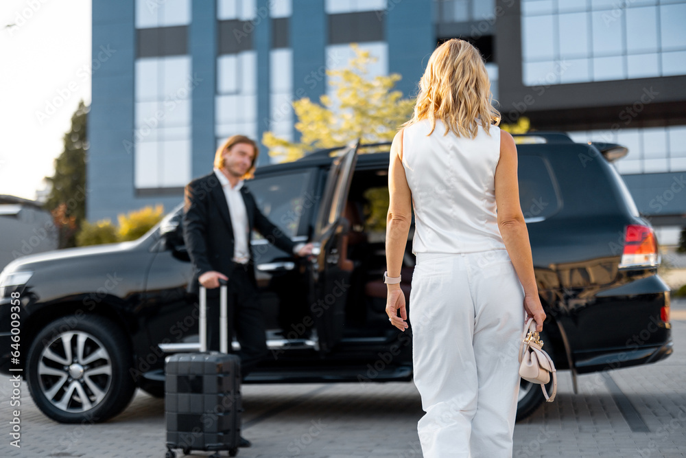 Wall mural elegant business woman in white casual clothes walks to the luxury car, man with a suitcase opens a 