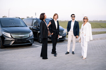 Group of an elegant business people have a conversation while standing together on a parking lot near vehicles outdoors. Colleagues and business partners talking during a break outdoors at dusk