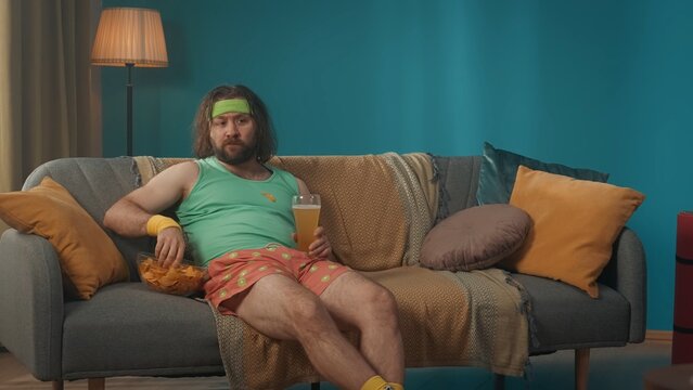 Obese middle-aged man with long hair sits in a room on a couch carelessly eating chips and drinking beer. The man is carefully watching a TV program on the television.