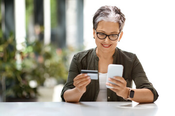 Cheerful attractive mature woman using bank card and phone