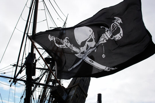 Pirate flag with skull and crossbones waving on a ship mast against a cloudy sky. A classic Jolly Roger flag flutters in the wind, symbolizing adventure and maritime history on a sailing vessel