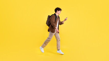 Fototapeta na wymiar Cheerful Student Guy Using Smartphone Walking With Backpack, Yellow Background