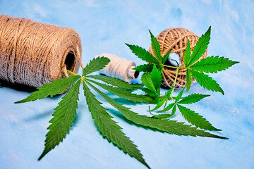 hemp leaf and branch lies next to spools of natural cannabis fibers, spools of thread on its side on a blue canvas and hemp seeds, close-up