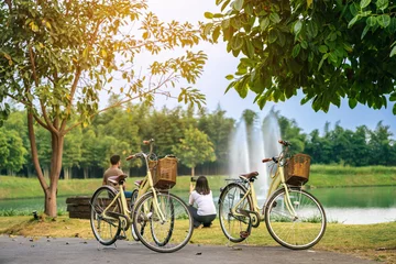 Deurstickers Two retro bicycles parked on the edge of a pond with happiness couple relaxing with pool and fountain in the background. Taking a rest after cycling in the park. Beautiful vintage bicycle in garden. © JinnaritT