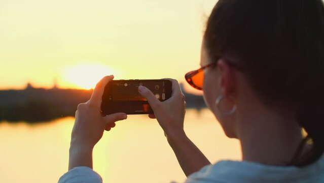 Woman takes photo of sunset sky with mobile phone. Beautiful girl takes pictures and videos of sunset on her smartphone. Idyllic nature landscape