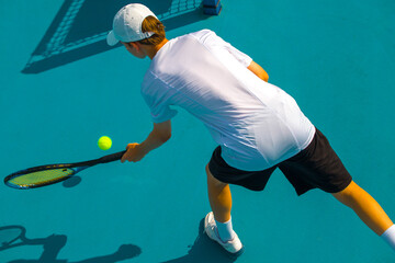 A young man plays tennis on a court with a hard blue surface on a summer sunny day