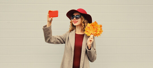 Autumn style outfit, stylish elegant happy woman posing taking selfie with mobile phone holds yellow maple leaves wearing hat, coat jacket and sunglasses on gray background