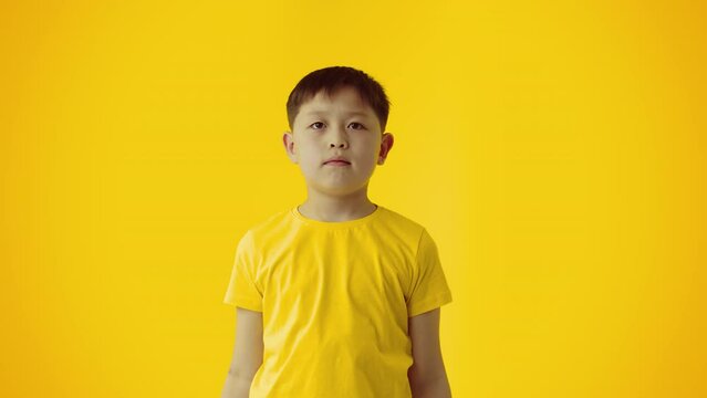 Young little asian boy looks extremely surprised and throws up his hands to the sides in bewilderment on yellow background. Concept of lack of child's understanding for further action