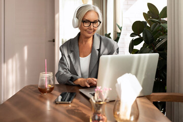 successful well-groomed middle-aged woman with glasses spends time at a laptop while sitting in a...